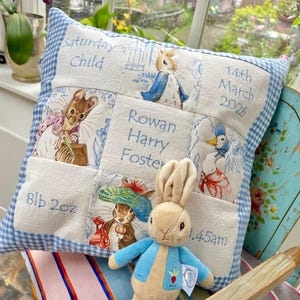 Può includere: Un cuscino quadrato patchwork bianco e blu con bordo a quadretti. Il cuscino presenta illustrazioni ricamate di personaggi delle fiabe e il testo "Saturday's Child", "Rowan Harry Foster", "14th March 2026" e "8lb 2oz". Un peluche di Peter Rabbit è davanti.