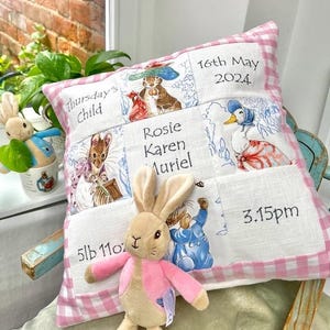 Può includere: Un cuscino per bambini personalizzato con un bordo a quadretti rosa. Il cuscino presenta illustrazioni ricamate di personaggi di Peter Rabbit e dettagli: "Thursday's Child", "16th May 2024", "Rosie Karen Muriel", "3.15pm" e "5lb 11oz".