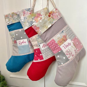May include: Three personalised Christmas stockings in blue, red, and grey, each with a floral patterned cuff. The stockings feature patchwork designs with polka dots and stars, and are labelled with names in embroidered script.