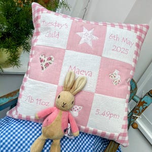 Può includere: Un cuscino quadrato con un bordo a quadretti vichy rosa e bianchi e una griglia di quadrati in tessuto bianco. Il cuscino presenta testi ricamati in rosa tra cui "Tuesday's Child", "6th May 2025", "Maeva", "7lb 11oz" e "5.49pm", insieme ad applicazioni di una stella, un cuore floreale e scarpine da neonato. Un coniglio di peluche beige con una giacca rosa è posizionato davanti.