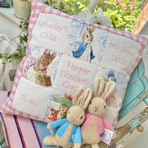 Può includere: Un cuscino decorativo quadrato con bordo a quadretti vichy rosa e bianco, con testo ricamato 'Saturday's Child', '6th June 2026', 'Harper Elizabeth Grace' e '7lb 8oz', illustrazioni di Peter Rabbit e due peluche a forma di coniglio.