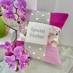 Pode incluir: Uma almofada de patchwork com um design floral e as palavras "Special Mother" bordadas em cinza sobre um quadrado branco. A almofada é feita de tecido rosa, cinza e branco.