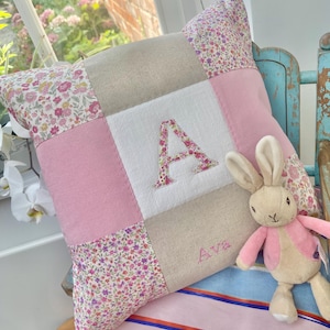 May include: A square patchwork cushion featuring a central white panel with a large floral-patterned letter A, surrounded by alternating solid pink and beige squares and floral-patterned fabric. The name Ava is embroidered in pink below the letter A.