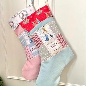 May include: Two Christmas stockings with patchwork designs. One is light pink, the other is light blue. Both have a Peter Rabbit theme with the names "Am" and "Alfie" embroidered on them. The stockings are hanging on a white door.