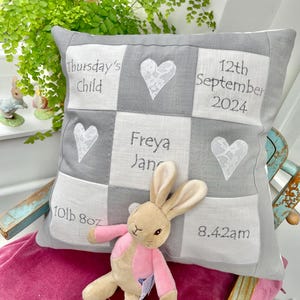 Può includere: Un cuscino a patchwork grigio e bianco con dettagli ricamati. Il cuscino presenta un cuore, il nome Freya Jane, la data 12 settembre 2024 e l'ora 8:42. Il cuscino include anche il testo "Thursday's Child" e "10lb 8oz". Un coniglietto di peluche rosa e marrone è seduto sul cuscino.