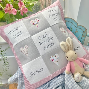 May include: A square decorative cushion with a pink polka dot border, featuring a grid of white and grey fabric squares. Embroidered text includes 'Sunday's Child', '16th August 2026', 'Everly Brooke Jones', and '6lb 8oz', accented by floral hearts.