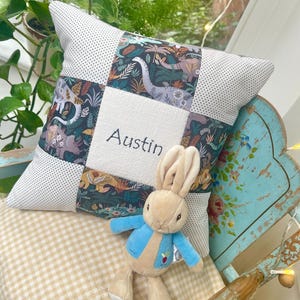 Op de afbeelding: Een vierkant patchwork kussen met een dinosaurusprint en de naam "Austin" geborduurd op een witte vierkant. Een knuffelkonijn met een blauw jasje ernaast.