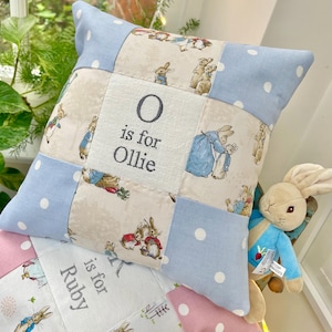Puede incluir: Un cojín decorativo cuadrado con un diseño de patchwork de tela azul claro con lunares blancos y tela beige estampada con ilustraciones de Peter Rabbit. El panel central está bordado con el texto O is for Ollie en gris oscuro.