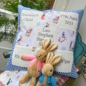 Può includere: Un cuscino per bambini personalizzato con bordo a quadretti blu e design patchwork. Il cuscino presenta dettagli ricamati, tra cui il nome "Leo Stephen", la data di nascita e il peso. Due peluche a forma di coniglietto sono davanti al cuscino.