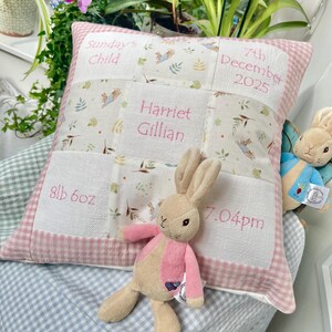 Può includere: Un cuscino personalizzato per bambini con bordo a quadretti rosa e dettagli ricamati. Il cuscino mostra il nome "Harriet Gillian", il peso alla nascita 3,8 kg, l'ora di nascita 19:04 e la data di nascita 7 dicembre 2025. Un peluche a forma di coniglietto è davanti al cuscino.