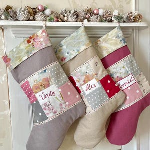 Könnte beinhalten: Drei personalisierte Weihnachtsstrümpfe mit floralen und gepunkteten Patchwork-Designs. Jeder Strumpf hat ein anderes Farbschema, mit Namen auf der Vorderseite gestickt. Die Strümpfe hängen an einem weißen Regal.