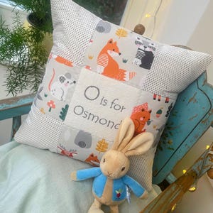 Op de afbeelding: Een patchwork kussen met dierenillustraties, waaronder een vos en een muis, en de tekst "O is for Osmond". Het kussen wordt vergezeld door een knuffelkonijn in een blauw jasje.