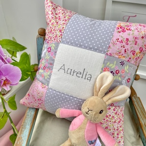 May include: A patchwork cushion embroidered with the name "Aurelia". The cushion features floral and polka dot patterns in pink, grey, and white. A stuffed bunny toy with pink clothing sits in front of the cushion, creating a soft, inviting scene.