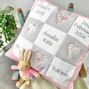 Può includere: Un cuscino patchwork quadrato con quadrati di tessuto bianchi e grigi alternati con testo ricamato tra cui "Friday's Child", "Amelie Kate", "2nd February 2024", "6lb" e "9.30pm", accentuato da applicazioni a cuore floreali e un bordo rosa.