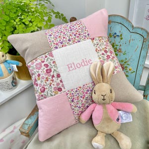 Peut inclure: Un coussin de patchwork rose et beige avec un motif floral et le nom "Elodie" brodé dessus. Un jouet en peluche de lapin beige est assis sur le coussin.