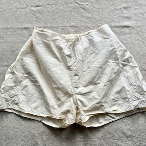 May include: Cream-colored vintage shorts with floral embroidery near the hem. The shorts are made of a lightweight fabric and have a high waist. The garment is laid flat on a textured, neutral-toned surface.