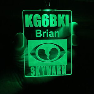 Könnte beinhalten: Ein leuchtend grünes rechteckiges Etikett mit dem Text "KG6BKI" über "Brian" und einer stilisierten Augengrafik. Unter der Grafik steht das Wort "SKYWARN". Das Etikett wird in einer Hand gehalten.