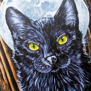 May include: A close-up of a black cat with bright yellow eyes. The cat is painted on a wooden surface with a white moon behind it. The painting is signed "Wildlife Artist Hayley".