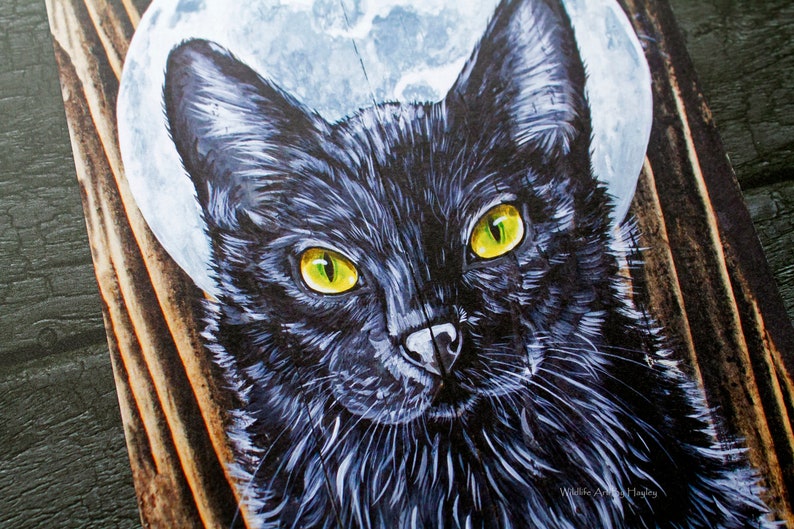 May include: A close-up of a black cat with bright yellow eyes. The cat is painted on a wooden surface with a white moon behind it. The painting is signed "Wildlife Artist Hayley".