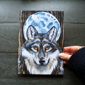 May include: A painting of a wolf with a full moon behind it. The wolf is gray and brown with yellow eyes. The moon is blue and white. The painting is on a wooden background.