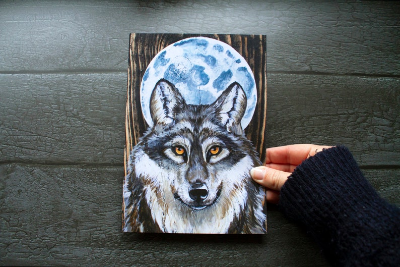 May include: A painting of a wolf with a full moon behind it. The wolf is gray and brown with yellow eyes. The moon is blue and white. The painting is on a wooden background.