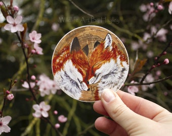 May include: A round sticker with a woodgrain background featuring a painting of two red foxes with their heads touching. The foxes are painted in a realistic style with white fur and brown accents. The sticker has the text "Wildlife Art By Hayley" printed at the top.