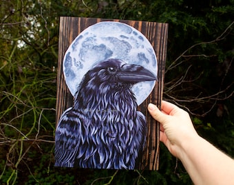 May include: A black raven with a blue-grey moon behind it, painted on a dark wood background.