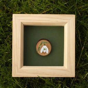 May include: A framed artwork depicting a snowdrop flower painted on a wooden slice. The artwork is set against a dark green background within a light-coloured wooden frame. The frame rests on a bed of green grass.