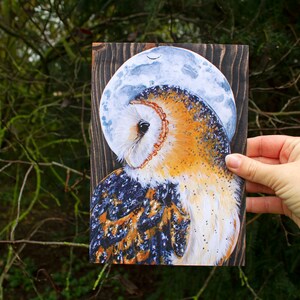 May include: A painting of a barn owl with a white and brown patterned design, perched against a dark wood background with a blue and white crescent moon behind it.