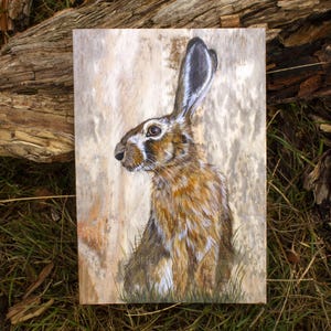 May include: A rectangular wooden panel featuring a detailed painting of a brown and white hare with long ears. The hare is depicted in a seated position, surrounded by subtle green grass at the base, set against a light, textured wood grain background.