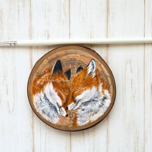 May include: A painting of two red foxes with closed eyes, cuddling on a wooden slice. The foxes are painted in a realistic style with detailed fur and features.