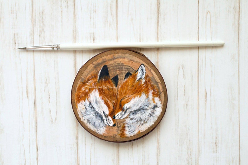 May include: A painting of two red foxes with closed eyes, cuddling on a wooden slice. The foxes are painted in a realistic style with detailed fur and features.