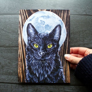 May include: A black cat with yellow eyes stares directly at the viewer. The cat is in front of a full moon and a woodgrain background.