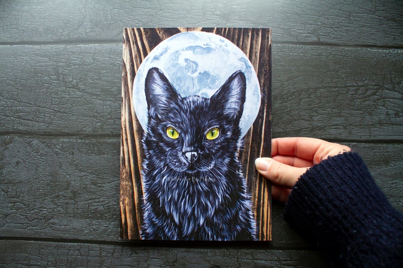 May include: A black cat with yellow eyes stares directly at the viewer. The cat is in front of a full moon and a woodgrain background.