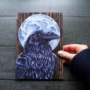 May include: A black raven with a blue-grey moon behind it. The raven is depicted in a realistic style with detailed feathers. The moon is a full moon with a textured surface. The image is printed on a wooden background.