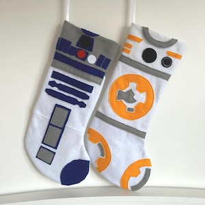 May include: Two white Christmas stockings with Star Wars-themed designs. One features a blue, gray, and white R2-D2 design, and the other has an orange, gray, and white BB-8 design. Both stockings are hanging against a white background.