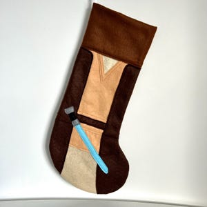 May include: A brown and tan Christmas stocking with a light blue lightsaber design. The stocking is made of felt and features a brown cuff and a tan and brown body. The lightsaber is light blue with a black and silver handle.