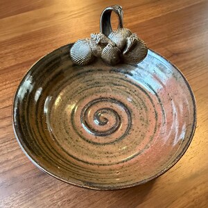 May include: A handmade ceramic bowl with a spiral design in the center. The bowl has a handle and is decorated with seashell accents. The colors are earthy tones of brown, tan, and a hint of pink.