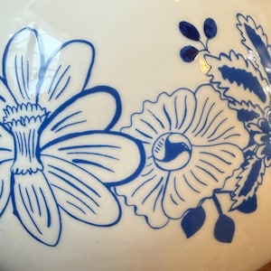 May include: A white ceramic vase with a blue floral design. The flowers are stylized with detailed petals and leaves. The vase has a smooth, glossy finish, and the blue color provides a striking contrast against the white background.