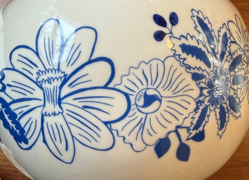 May include: A white ceramic vase with a blue floral design. The flowers are stylized with detailed petals and leaves. The vase has a smooth, glossy finish, and the blue color provides a striking contrast against the white background.