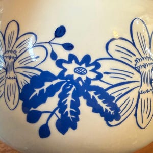 May include: A white ceramic vase with a glossy finish, adorned with a repeating pattern of large blue floral designs. The flowers have detailed petals and are arranged in a stylized manner, creating a decorative aesthetic.