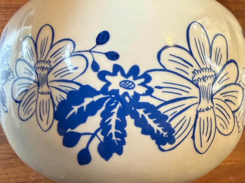 May include: A white ceramic vase with a glossy finish, adorned with a repeating pattern of large blue floral designs. The flowers have detailed petals and are arranged in a stylized manner, creating a decorative aesthetic.