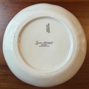 May include: A white ceramic bowl with a slightly irregular rim. The bottom of the bowl features the text "Lauren McIntosh exclusively for ANTHROPOLOGIE" and "MADE IN CHINA / FABRIQU&Eacute; EN CHINE". The bowl is on a wooden surface.