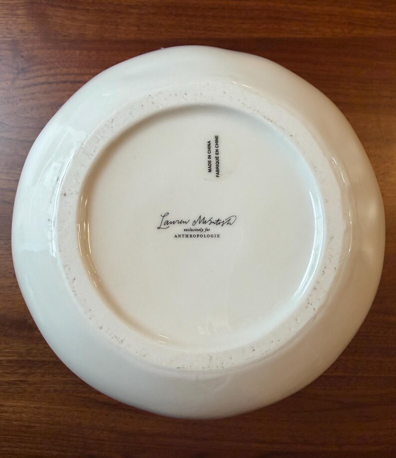 May include: A white ceramic bowl with a slightly irregular rim. The bottom of the bowl features the text "Lauren McIntosh exclusively for ANTHROPOLOGIE" and "MADE IN CHINA / FABRIQU&Eacute; EN CHINE". The bowl is on a wooden surface.