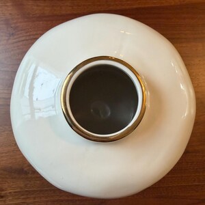 May include: A cream-colored ceramic vase with a wide, rounded body and a gold-colored rim around the opening. The interior is dark. The vase is set on a wooden surface.