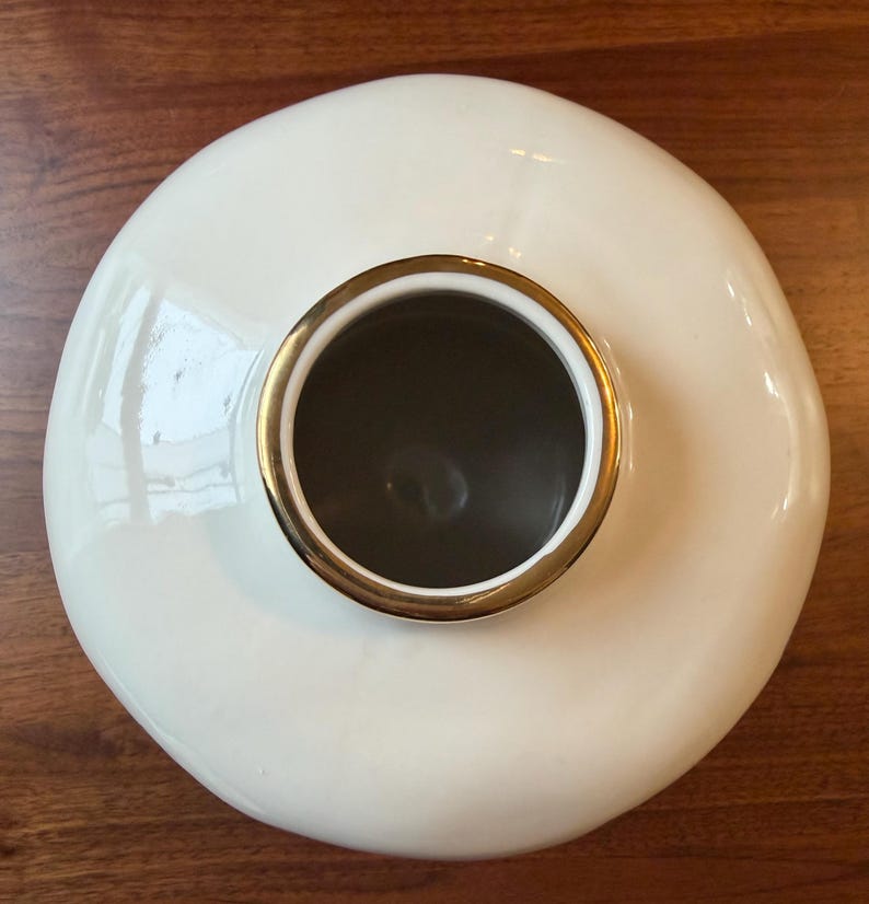 May include: A cream-colored ceramic vase with a wide, rounded body and a gold-colored rim around the opening. The interior is dark. The vase is set on a wooden surface.