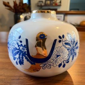 May include: A white ceramic vase with a gold rim and a gold bird design. The vase features blue floral patterns and a gold accent on the bird. The vase is a decorative item.