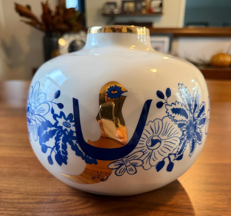 May include: A white ceramic vase with a gold rim and a gold bird design. The vase features blue floral patterns and a gold accent on the bird. The vase is a decorative item.