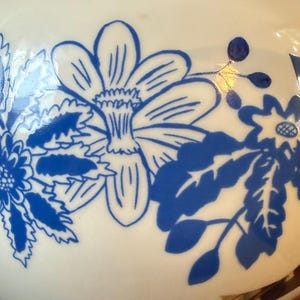May include: A white ceramic vase with a glossy finish, adorned with a blue floral pattern. The design features stylized flowers and foliage, creating a decorative and elegant aesthetic. The vase has a rounded shape.