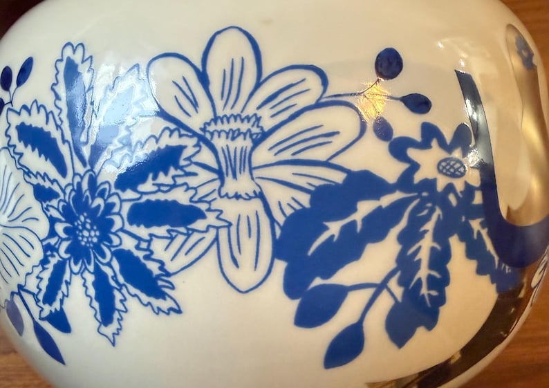 May include: A white ceramic vase with a glossy finish, adorned with a blue floral pattern. The design features stylized flowers and foliage, creating a decorative and elegant aesthetic. The vase has a rounded shape.